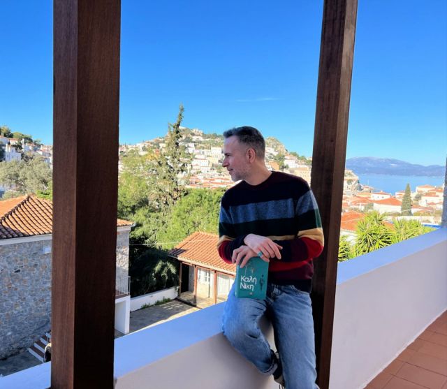 Ο Χρήστος Δασκαλάκης σε ένα μπαλκόνι κρατώντας το βιβλίο "Καλή Νίκη"